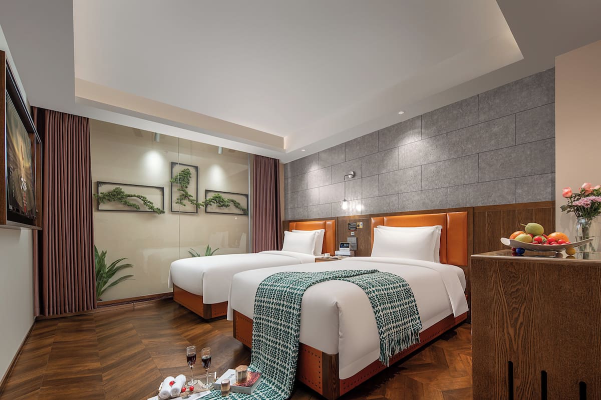 Habitación de hotel con dos camas, cabecera de madera, mesita de noche con frutero y televisor de pared.