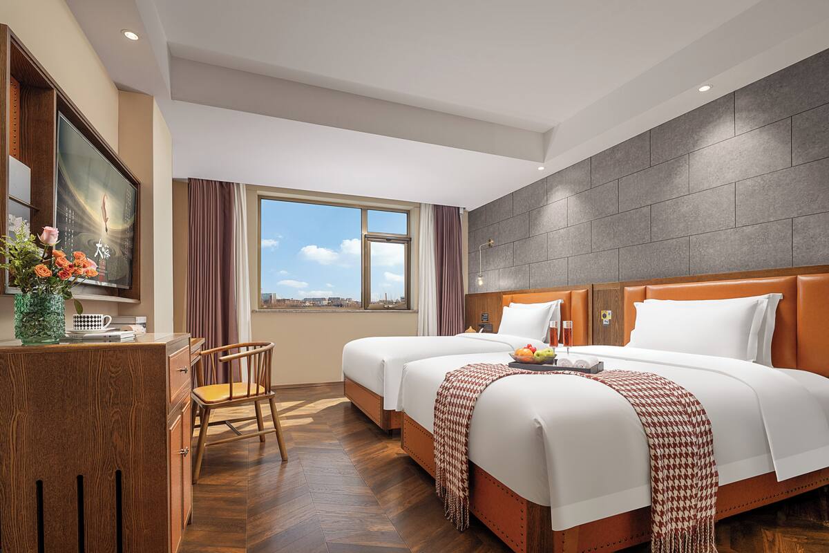 Habitación de hotel con dos camas, un escritorio con silla, un ventanal y vistas a la ciudad.