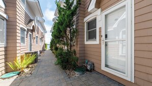 A narrow pathway between two buildings with a garage door, a small tree, and a utility box. - Hampton Bays Townhouse - Pool, Beach Access (Hampton Bays)