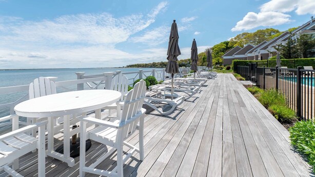 A wooden deck with white chairs and tables, a pool, and a view of the water. - Hampton Bays Townhouse - Pool, Beach Access (Hampton Bays)