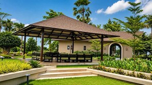 Un espace extérieur abrité, doté de bancs, entouré de jardins bien entretenus et de palmiers.