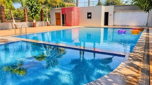 Una piscina rectangular con agua azul cristalina, rodeada por una terraza de baldosas y un pequeño edificio con techo rojo.