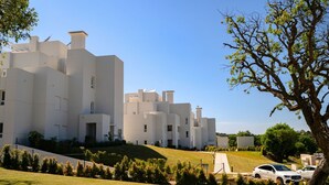Un edificio moderno blanco con varios balcones, rodeado de vegetación y coches aparcados.