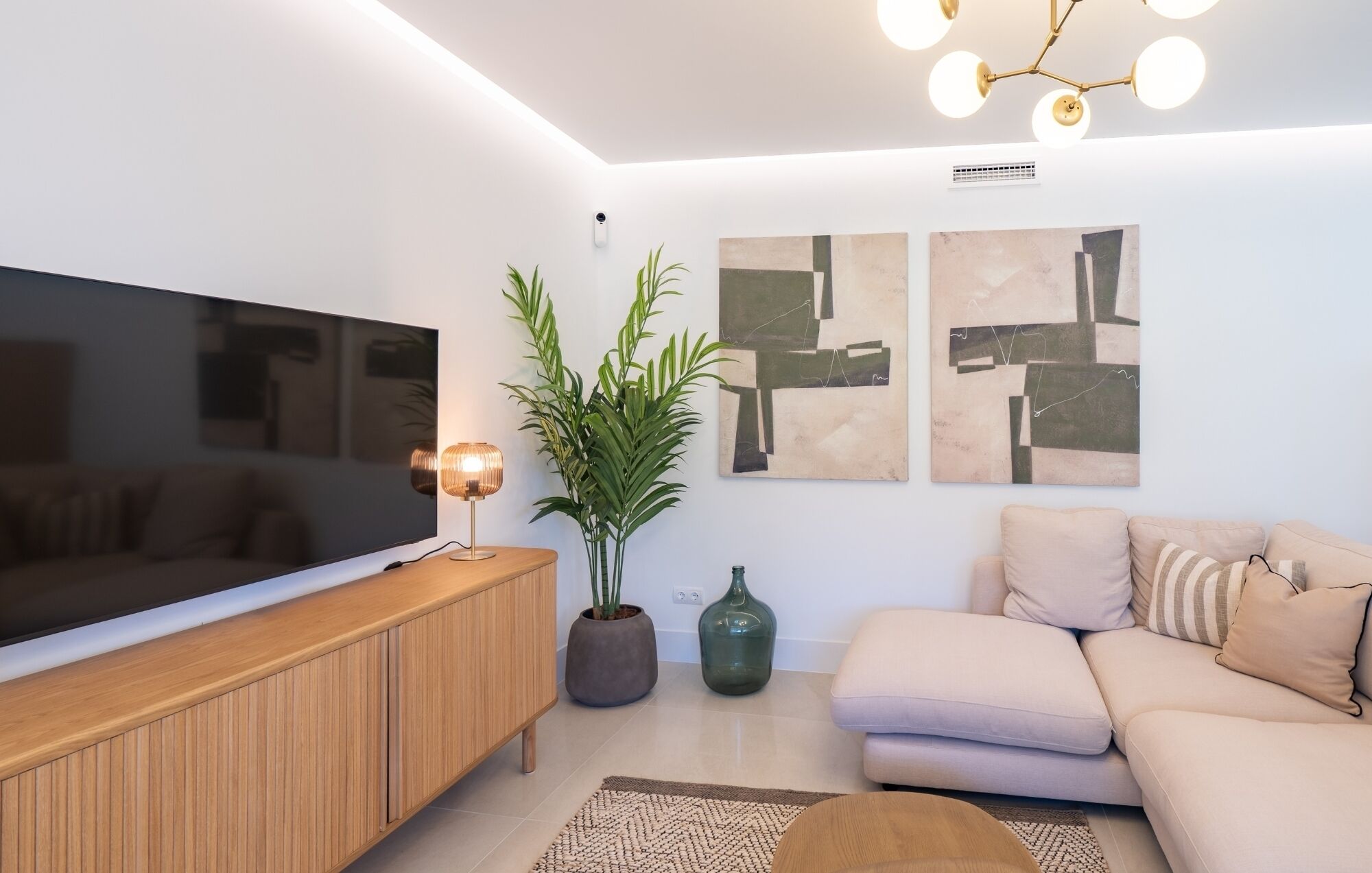 Un salon moderne avec un téléviseur à écran plat, un meuble en bois, une plante verte, une œuvre d’art abstraite et un canapé beige.