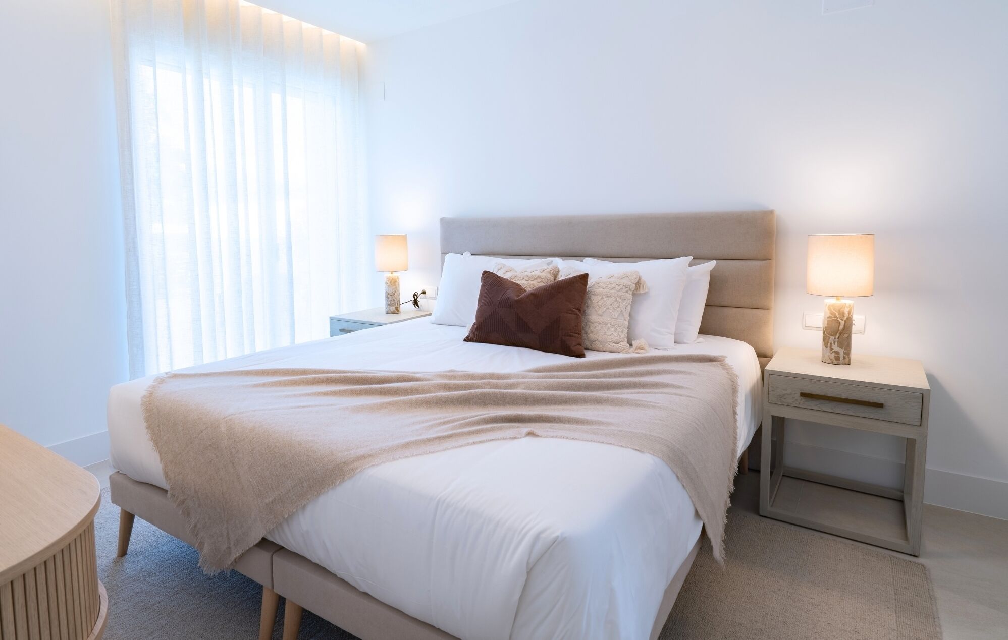 Een netjes opgemaakt bed met een hoofdbord, twee nachtkastjes met lampen, en een raam met dunne gordijnen.