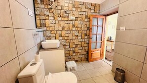 Une salle de bain avec un mur en pierre, des toilettes, un lavabo, un petit tabouret et une porte donnant sur une autre pièce.