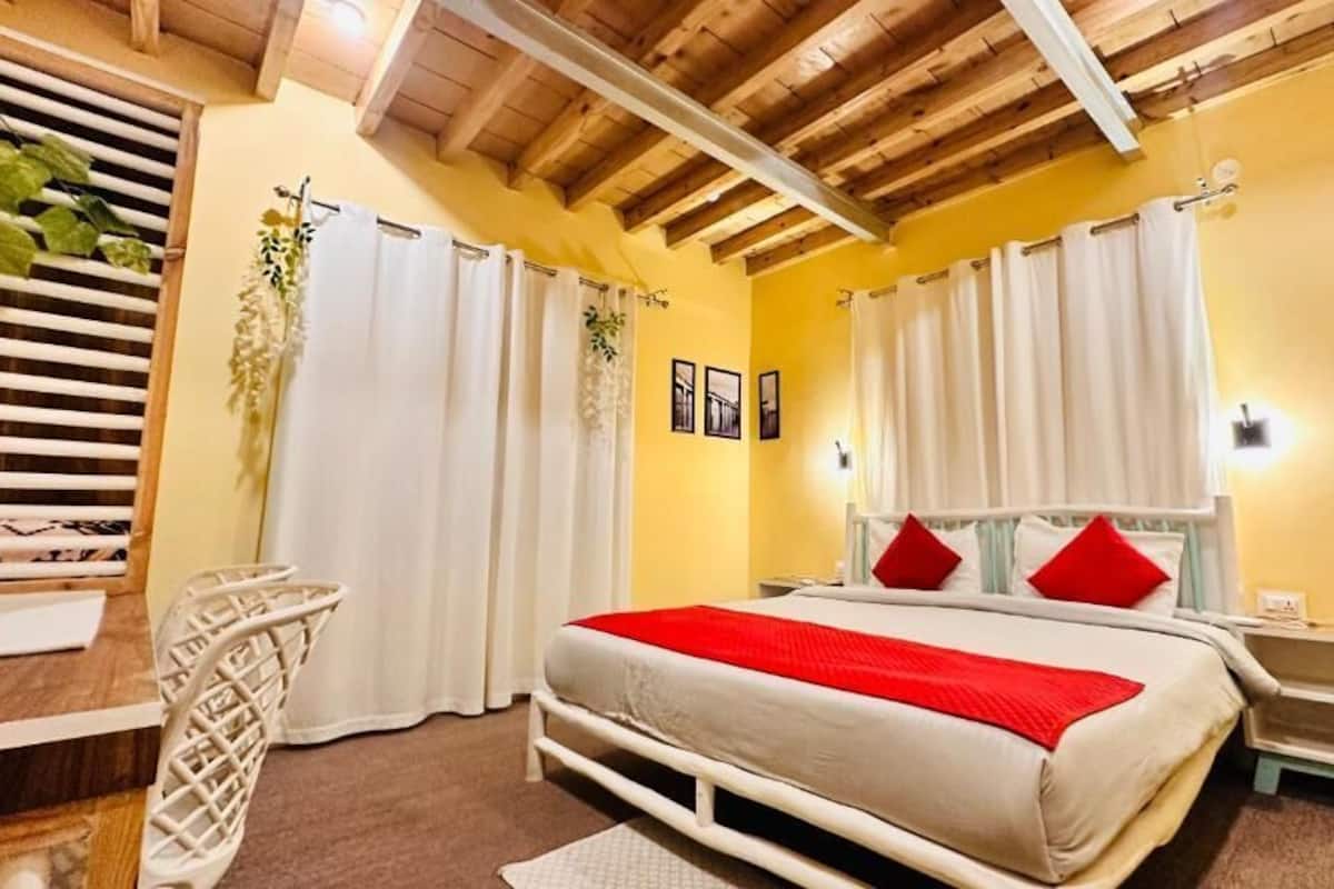 A bedroom with a bed, white curtains, a bedside table, a nightstand, a wooden ceiling, and a window with blinds.