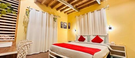 A bedroom with a bed, white curtains, a bedside table, a nightstand, a wooden ceiling, and a window with blinds.