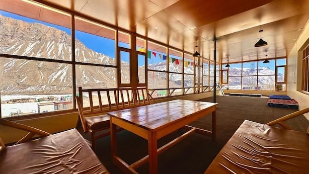 Une pièce avec une table et des bancs en bois, de grandes fenêtres donnant sur la montagne et des drapeaux aux couleurs vives.