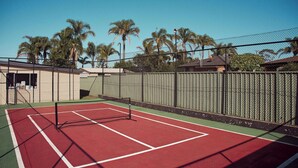 Ein Außen-Tennisplatz mit Netz, umgeben von einem Maschendrahtzaun, Palmen und Gebäuden im Hintergrund.