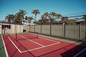 An outdoor tennis court with a net, surrounded by a chain-link fence, palm trees, and buildings in the background. - Yerriyong 3 - Family Cabin Sleeps 6 Mollymook (Mollymook)