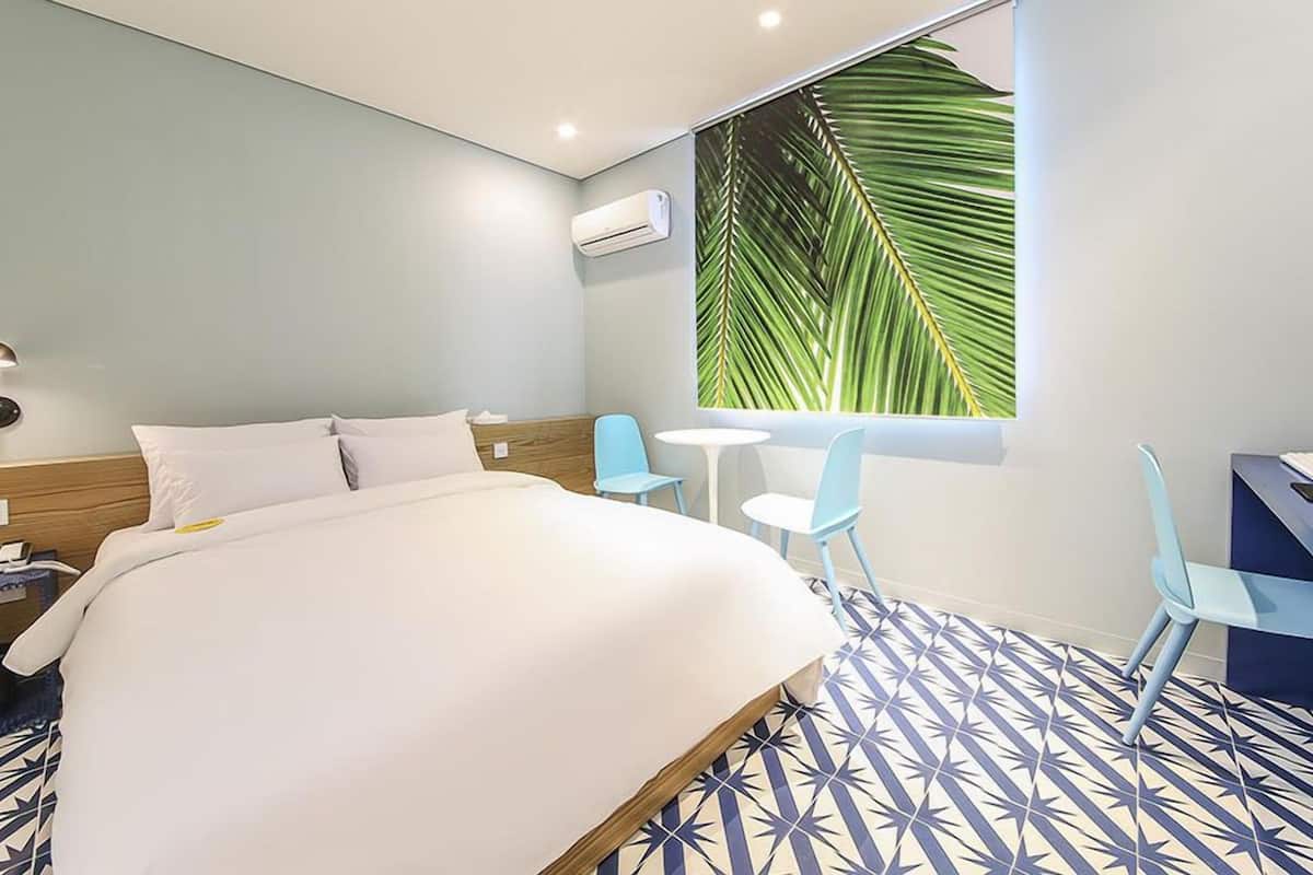 Une chambre d’hôtel comprenant un lit, un bureau, deux chaises, une petite table, un climatiseur fixé au mur et une grande fenêtre donnant sur un paysage de palmiers.