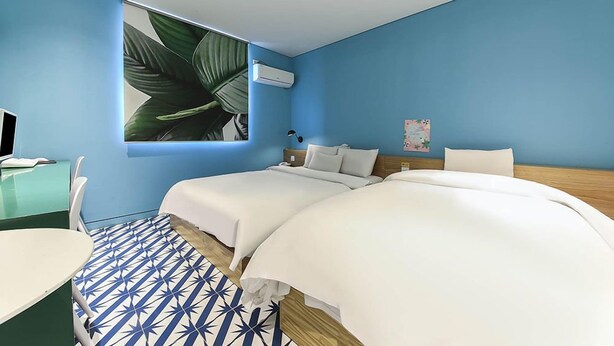 Hotellihuone, jossa on kaksi sänkyä, työpöytä tietokoneella, seinälle asennettu ilmastointilaite ja suuri ikkuna, jonka edessä on kasvi.