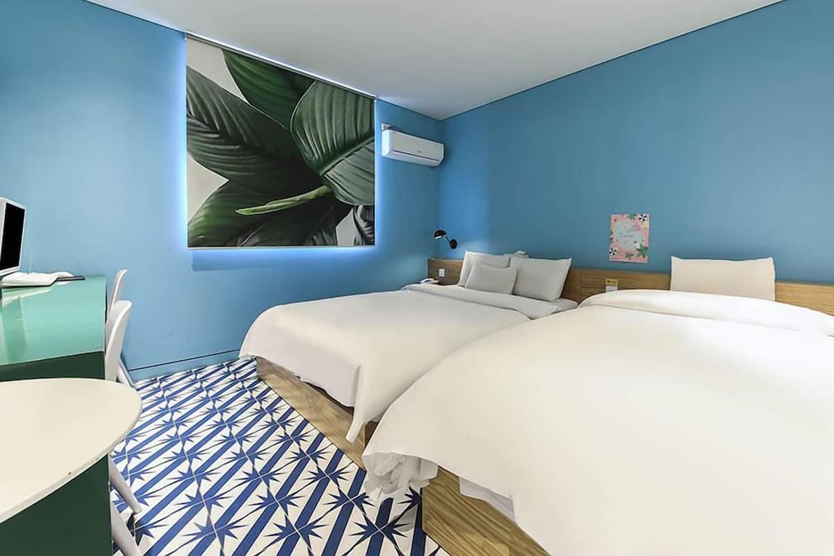 Une chambre d’hôtel avec deux lits, un bureau avec un ordinateur, un climatiseur fixé au mur et une grande fenêtre avec une plante.