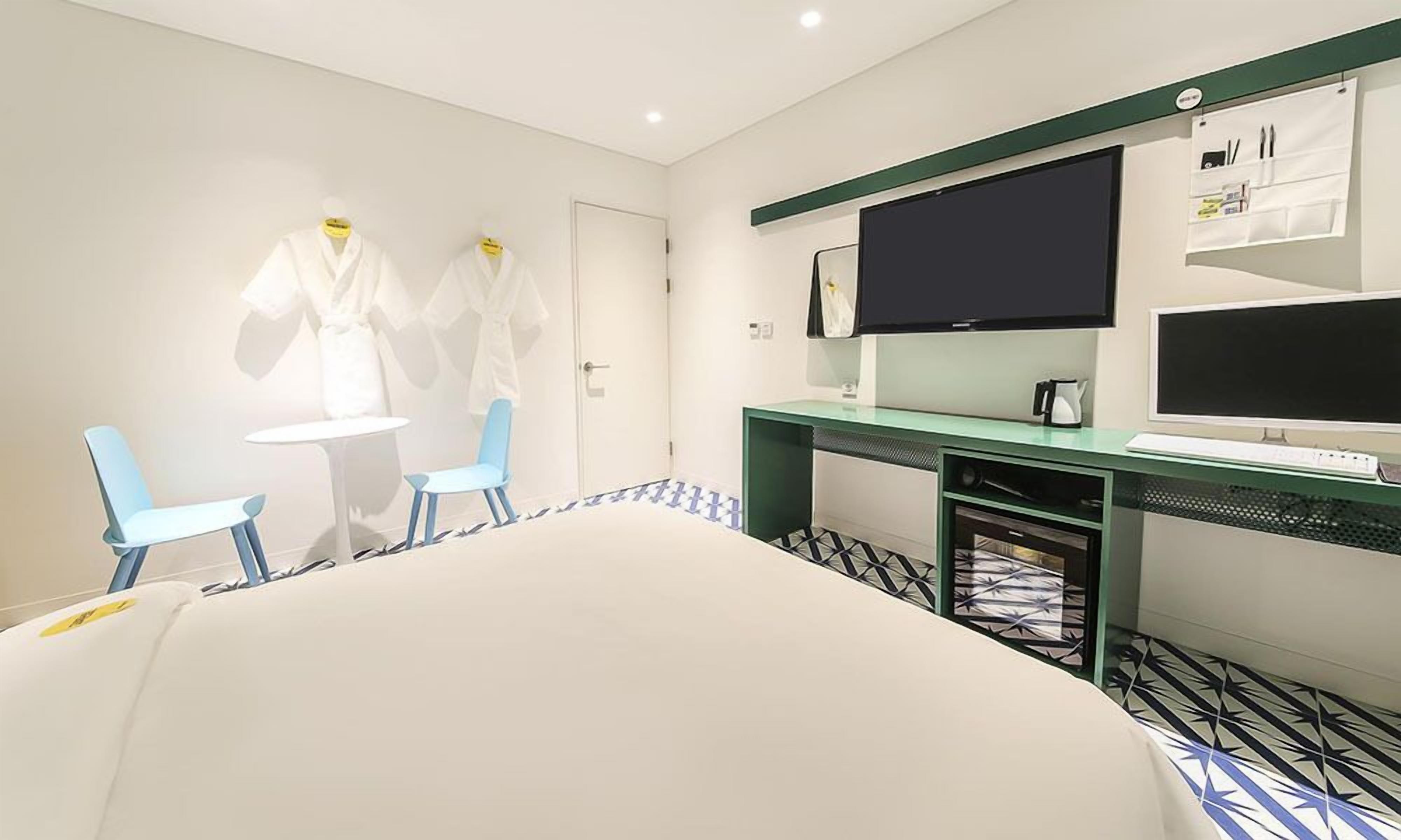 A hotel room with a bed, two chairs, a desk with a computer, a TV, and a small kitchenette.