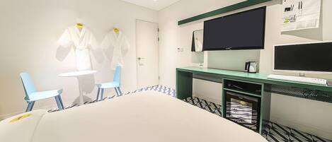 A hotel room with a bed, two chairs, a desk with a computer, a TV, and a small kitchenette.