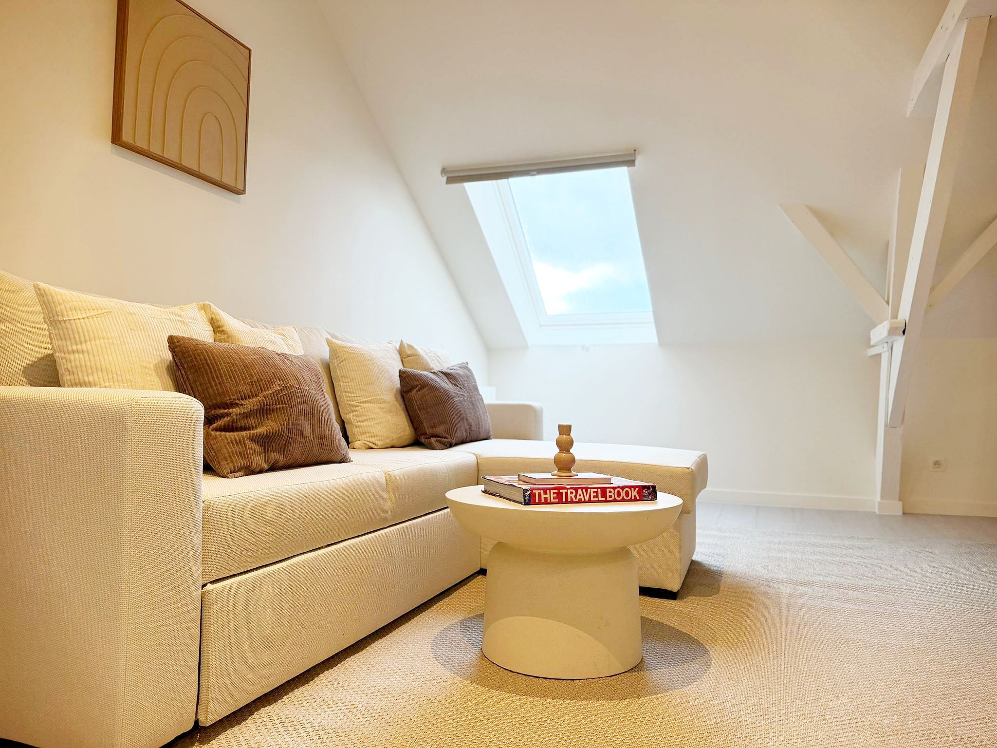 Une chambre à coucher confortable, aménagée dans un espace de type « loft », avec un plafond haut, un plafond en pente, un puits de lumière, un canapé blanc, une table basse blanche et une petite table de chevet.