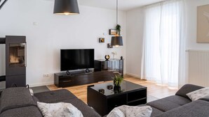 Ein modernes Wohnzimmer mit einem Flachbildfernseher, dunklen Möbeln und einem großen Fenster mit durchsichtigen Vorhängen.