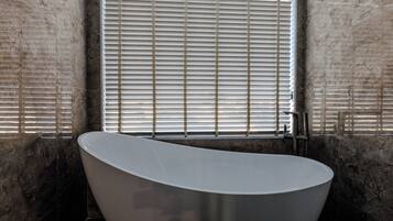 A modern bathroom with a freestanding bathtub, a window with blinds, and a textured wall.