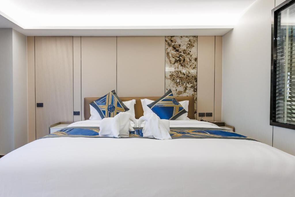 A hotel room with a large bed, a headboard with decorative pillows, a wall with a textured pattern, and a window with blinds.