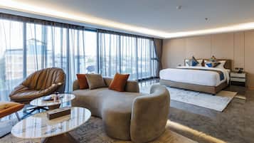 A modern hotel room with a large bed, a sofa, a leather armchair, a glass coffee table, and a view of the city through large windows.