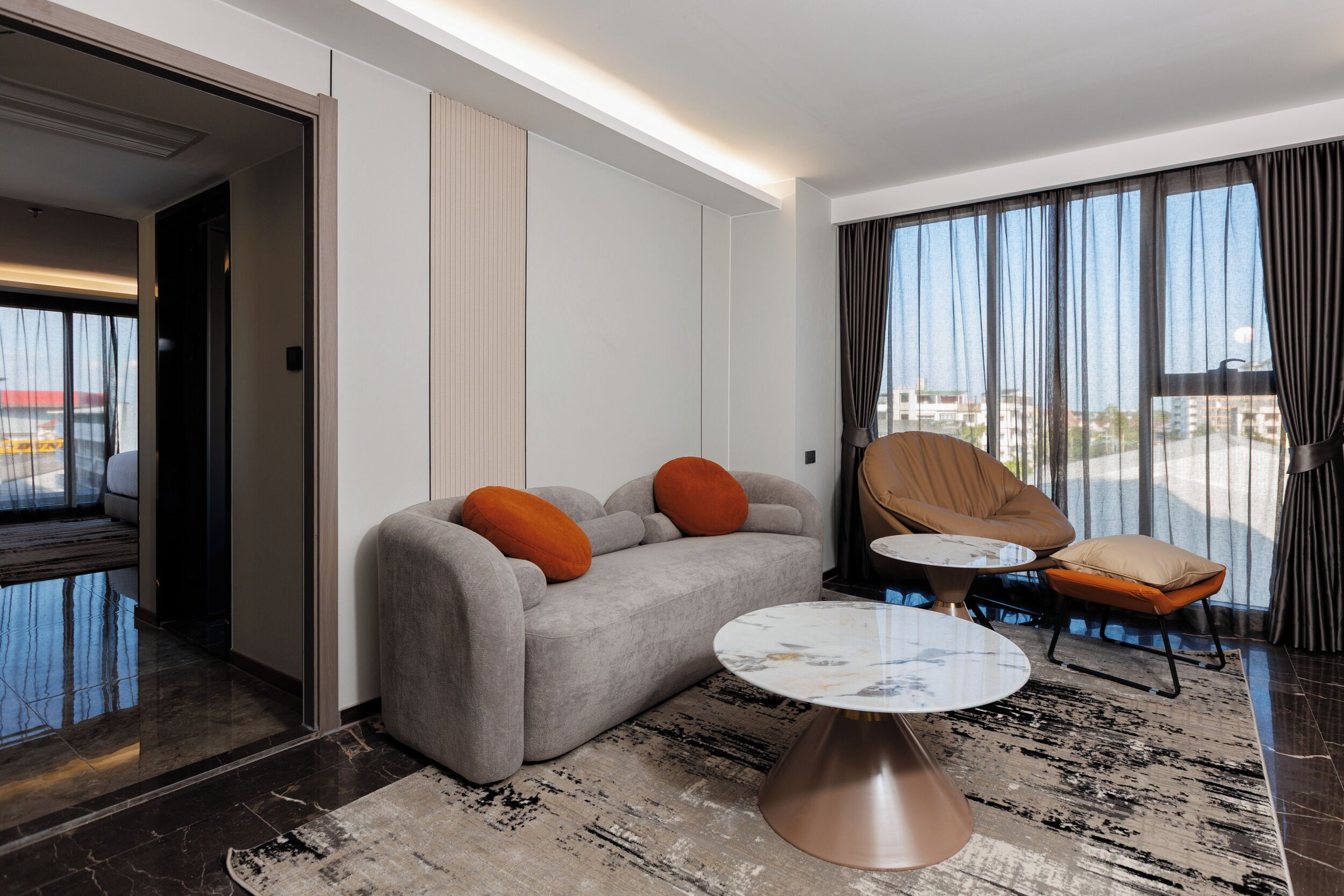 A modern hotel room with a grey sofa, two orange cushions, a round coffee table, a chair, and a large window with curtains.
