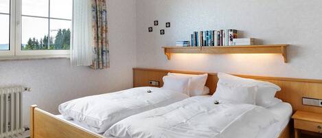 Een tweepersoonsbed met wit beddengoed, een houten hoofdbord, een nachtkastje, een plank met boeken, een raam met gordijnen en een radiator.