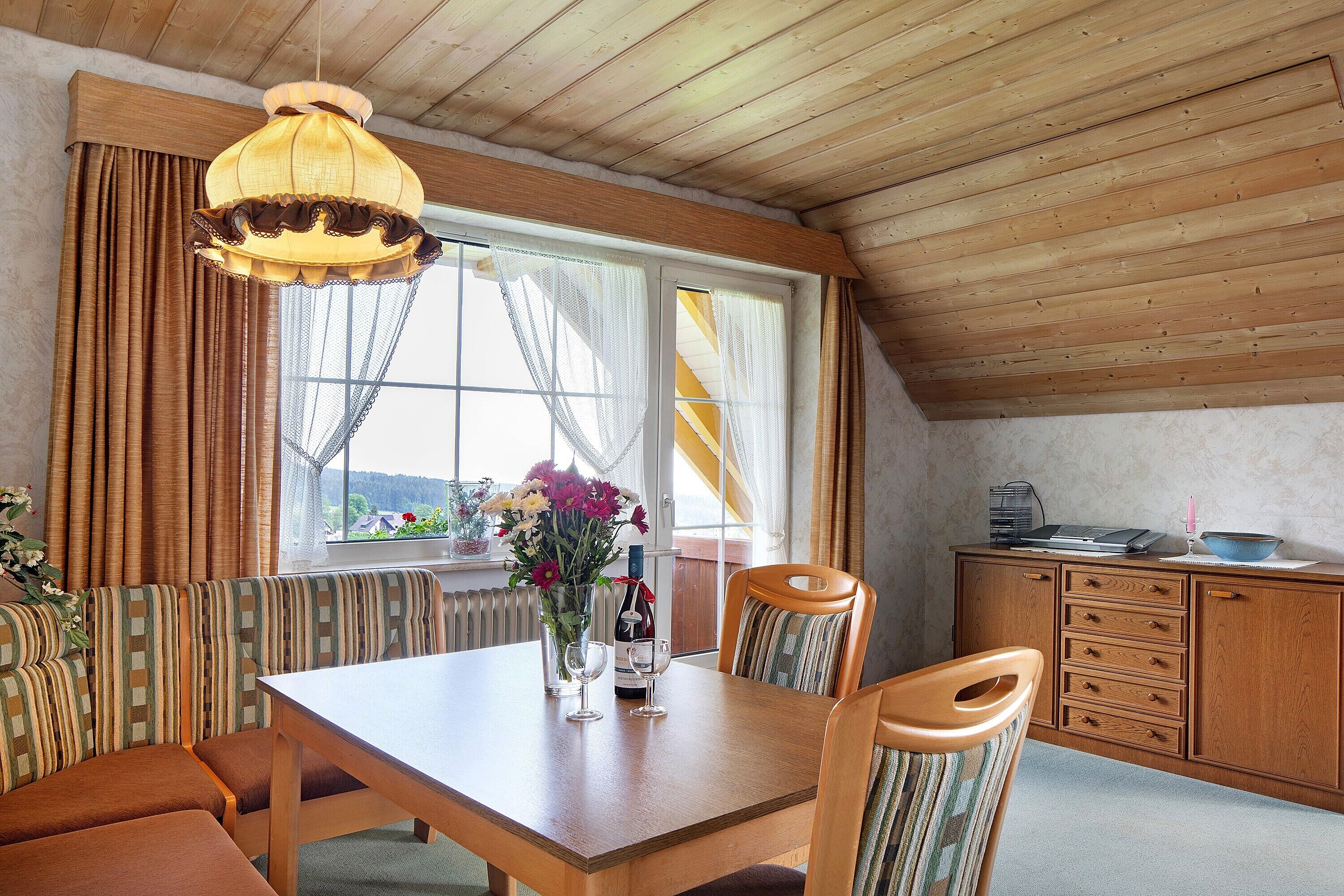 A dining area with a wooden table, chairs, a bench, a hanging light fixture, and a view through a window with curtains.