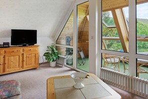 Ein Zimmer mit Fernseher auf einer Holzhocker, einem weißen Heizkörper, einem Esstisch mit weißer Schüssel und großen Fenstern mit Blick auf Grünfläche.