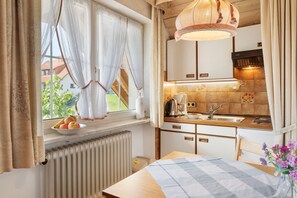 Eine kleine Küche mit Fenster, Heizkörper, Tisch mit kariertem Tischläufer und Blumen in einer Vase.
