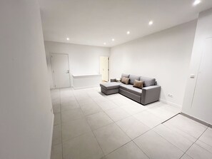 A modern, minimalist living room with a grey sectional sofa, a white bench, and a large window. - Central 1BR With 1LR Apt Great Location (Ghent)