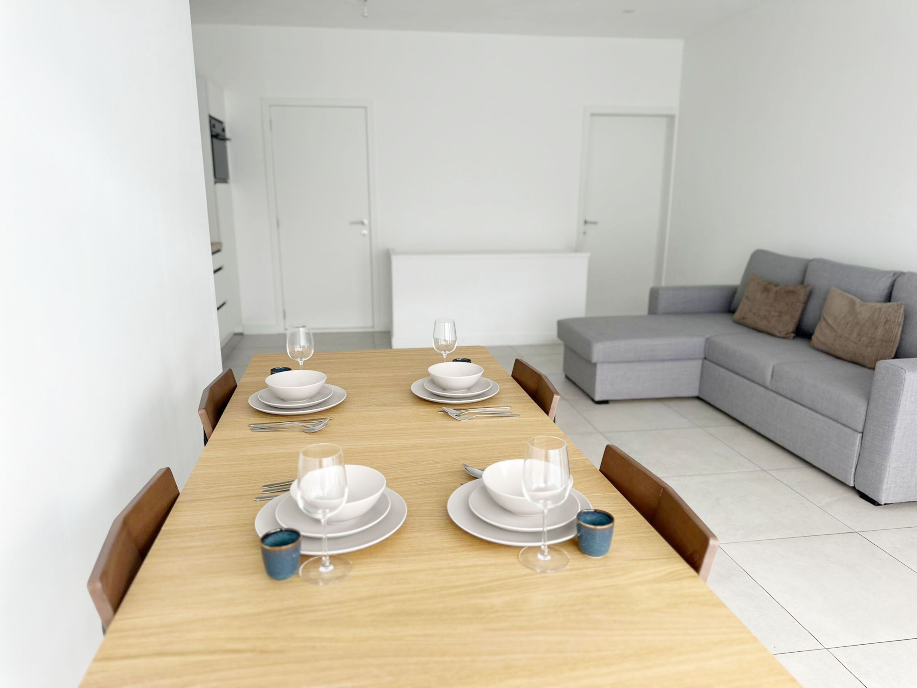 Un coin repas avec une table en bois dressée pour quatre personnes, un canapé gris et des murs blancs.