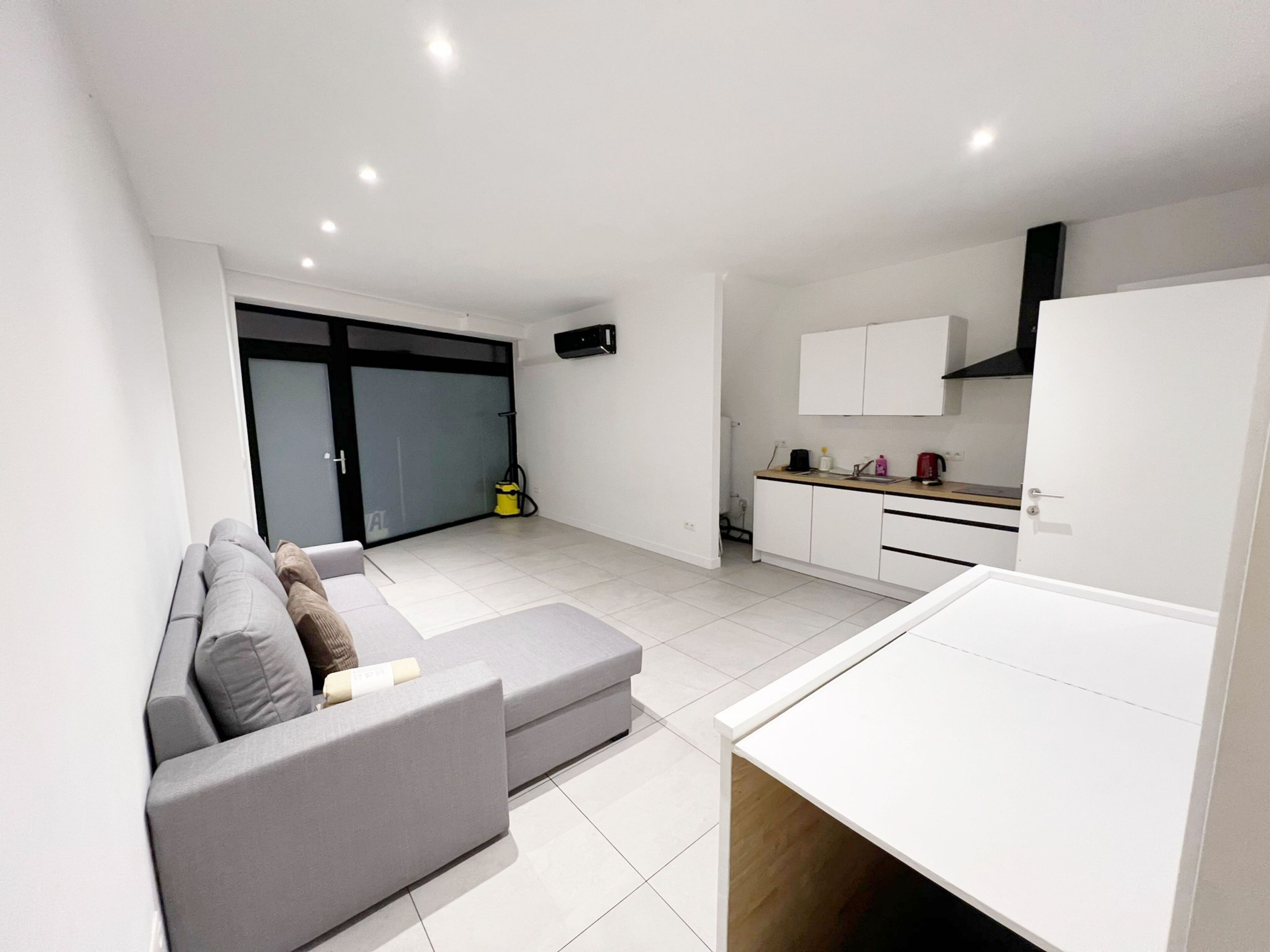 A modern living room with a grey sofa, a small dining table, and a kitchen area with white cabinets and a black range hood.