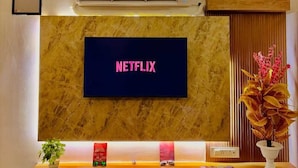A modern living room with a mounted TV displaying 'NETFLIX,' a ceiling fan, an air conditioning unit, and a wooden shelf with decorative items.