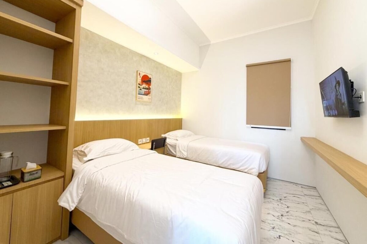 A hotel room with two beds, a wooden wardrobe, a small table with a lamp, a poster on the wall, and a window.
