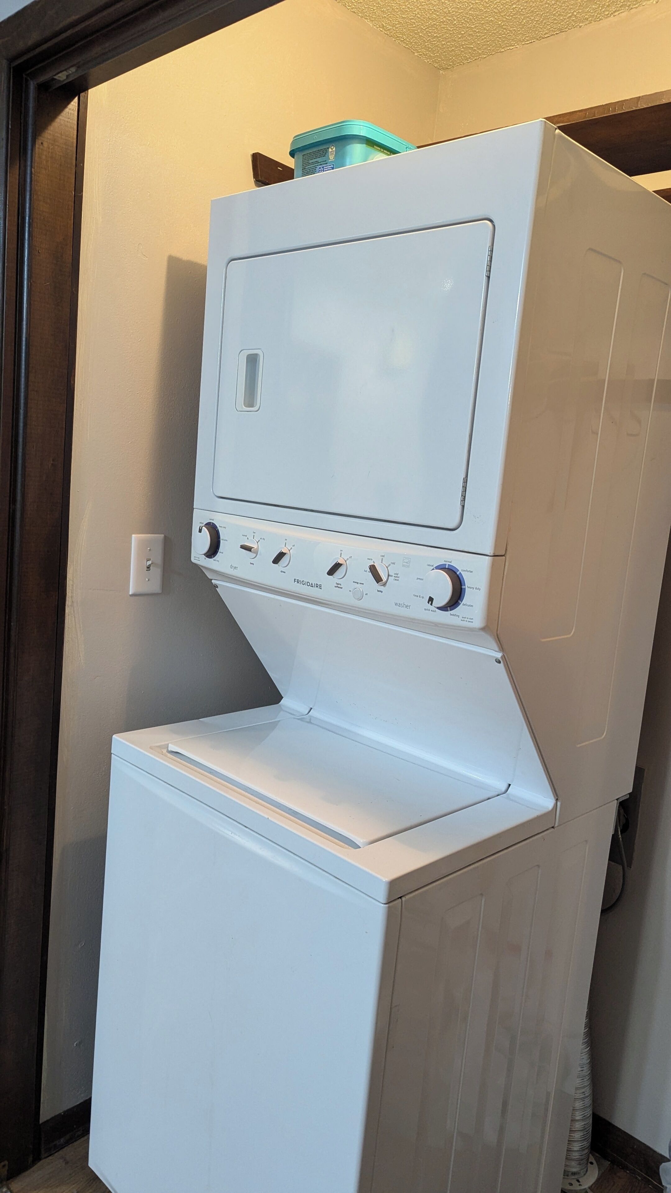 Un appareil compact combinant lave-linge et sèche-linge, doté d’un panneau de commande, installé dans un coin d’une pièce dont le sol est recouvert de parquet.