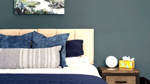 Una cama bien hecha con edredón azul y blanco, cabecera de madera y mesita de noche con caja de pañuelos y un reloj redondo.