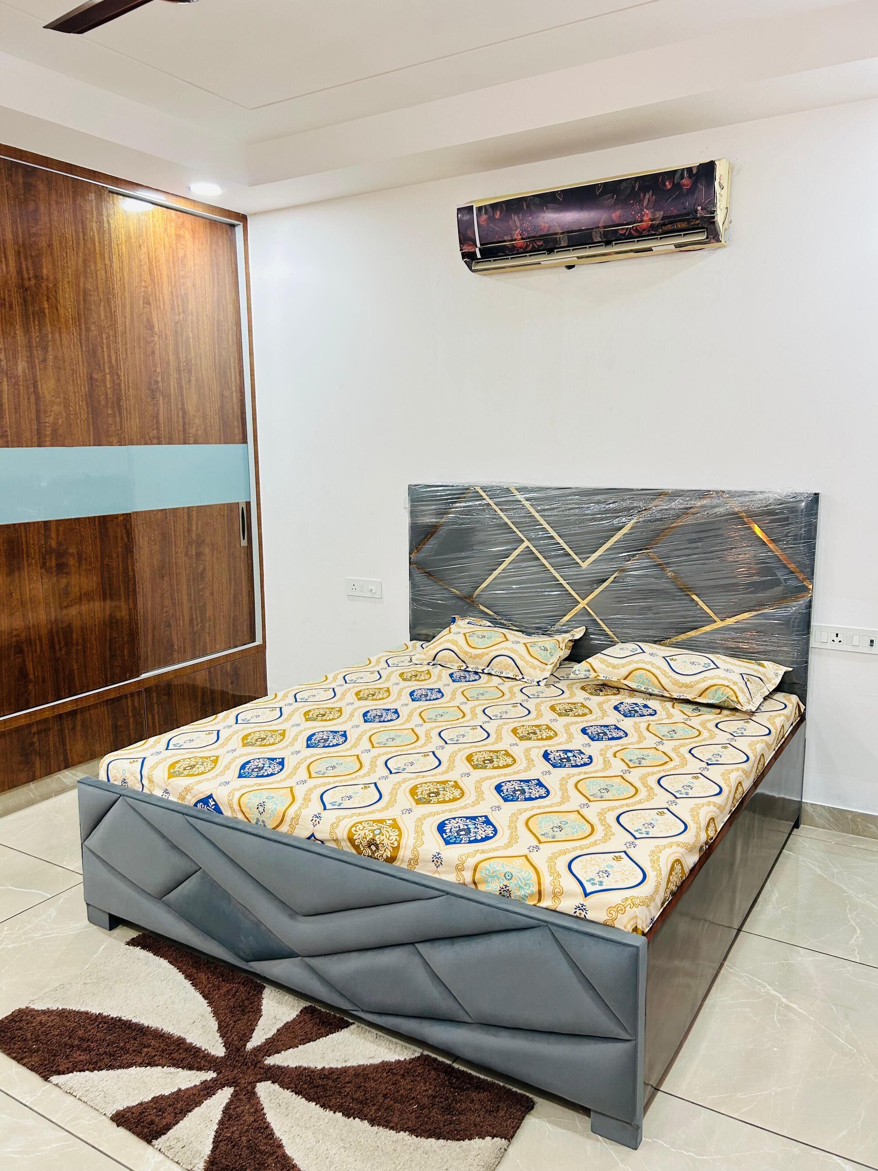 Ein Schlafzimmer mit einem Bett, einem Kopfteil, einem Holzschrank, einer Klimaanlage und einer gemusterten Bettdecke.