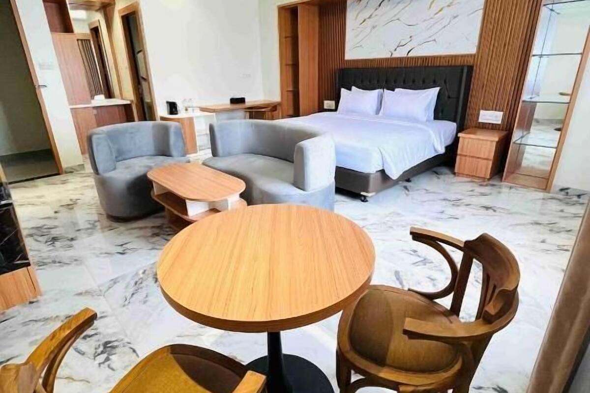A modern hotel room with a bed, two sofas, a round wooden table, and two wooden chairs.