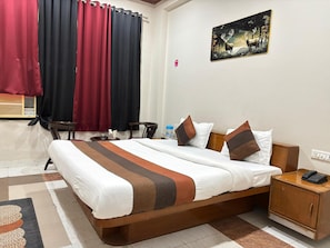 A hotel room with a bed, bedside table, nightstand, and a painting on the wall. - Hotel Dhola Maru (Kota)