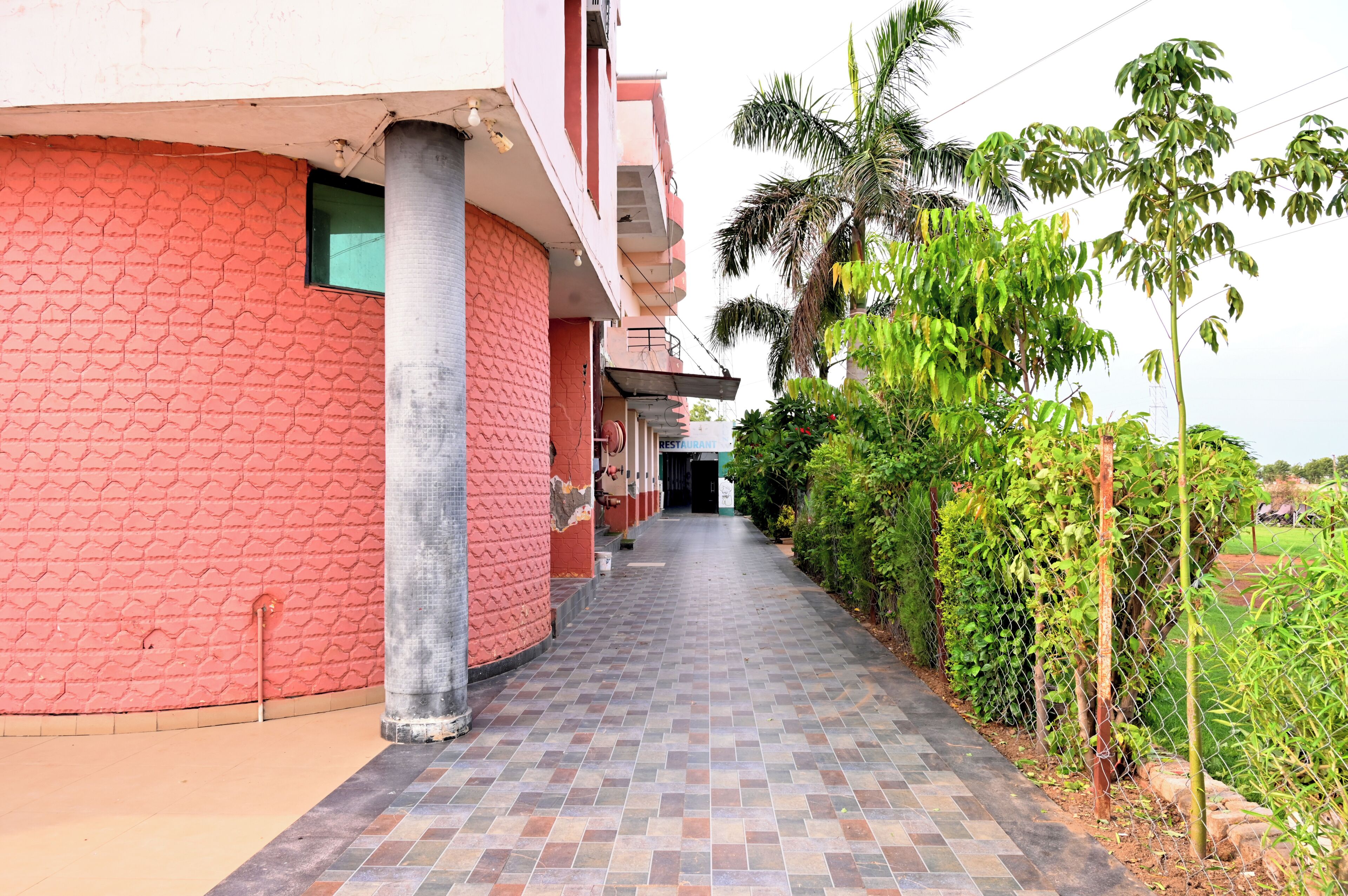A corridor with a tiled floor leading to a building entrance, surrounded by greenery and a chain-link fence.