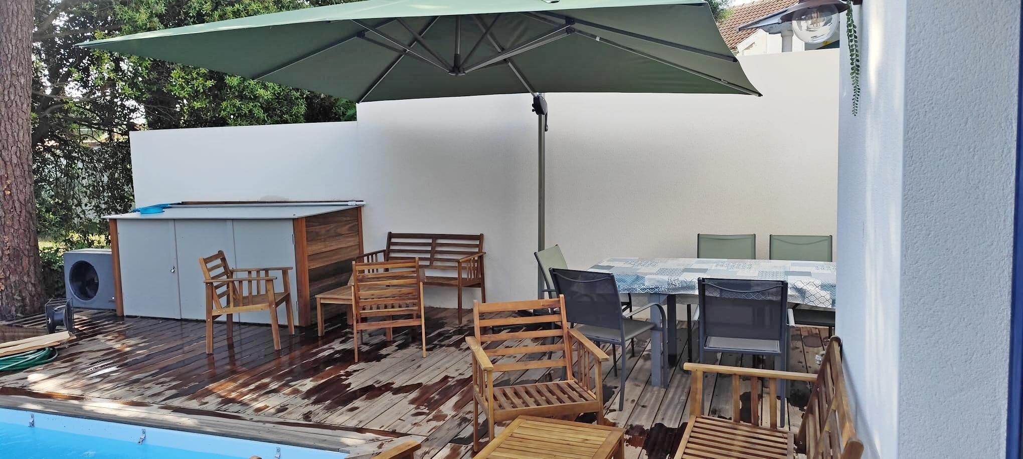 Una terraza de madera con mobiliario exterior, piscina y un espacio cubierto con mesa y sillas.