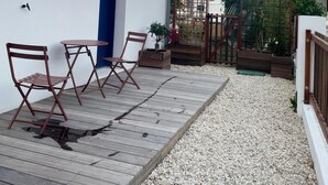Eine Holzterrasse mit Gartenmöbeln, ein weißes Gebäude mit blauen Fensterläden und ein Gartenbereich mit Bäumen und einem Holzzaun.