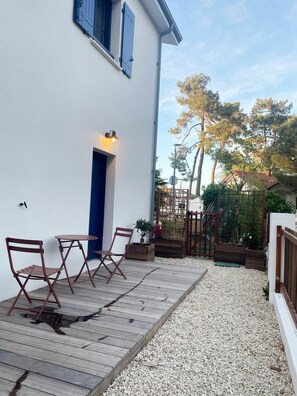 Eine Holzterrasse mit Gartenmöbeln, ein weißes Gebäude mit blauen Fensterläden und ein Gartenbereich mit Bäumen und einem Holzzaun.