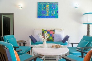 Ein Zimmer mit petrolfarbenen Stühlen, einem weißen Tisch, einem farbenfrohen gesteppten Sofa und einem Wandbild.