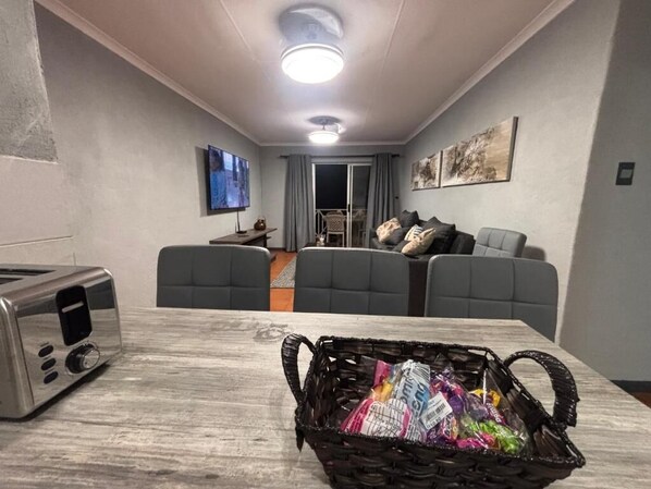 Una habitación con una mesa de comedor, un tostador, una cesta de aperitivos, un sofá y un televisor.