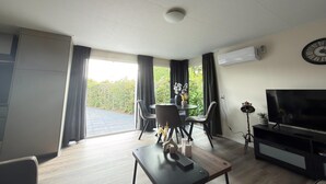 Ein modernes Wohnzimmer mit einem Flachbildfernseher, einem Esstisch mit Stühlen, einer Decken-Klimaanlage und einem großen Fenster mit Vorhängen.