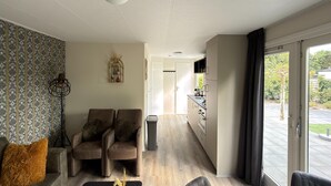 Ein modernes Wohnzimmer mit einer Couch, Sesseln, einem kleinen Tisch und einer Zimmerpflanze. Ein Flur führt zu einer Küche mit Fenster und Blick ins Freie.