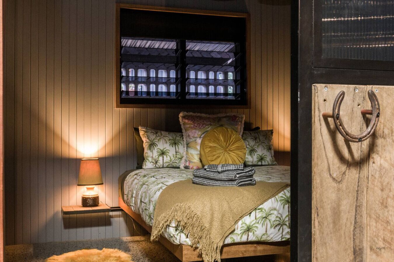 A bedroom with a bed, a nightstand, a lamp, a window with a view, and a wooden door.