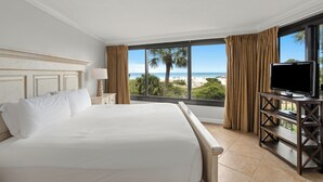 Habitación de hotel con una cama grande, un televisor sobre un soporte de madera y vistas a la playa a través de amplios ventanales.